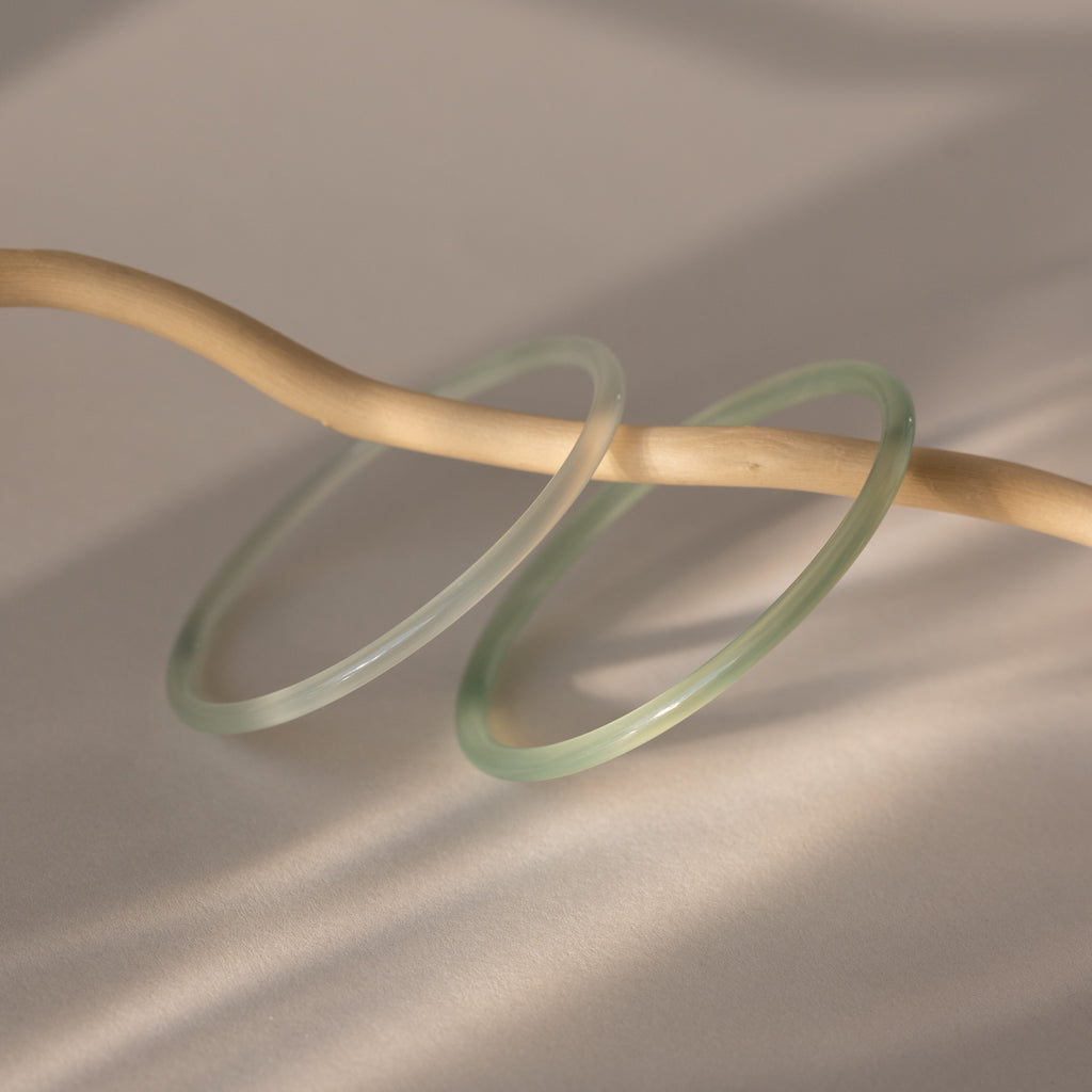 Set of two thin jade bangles in soft translucent green tones, displayed on a wooden stick.