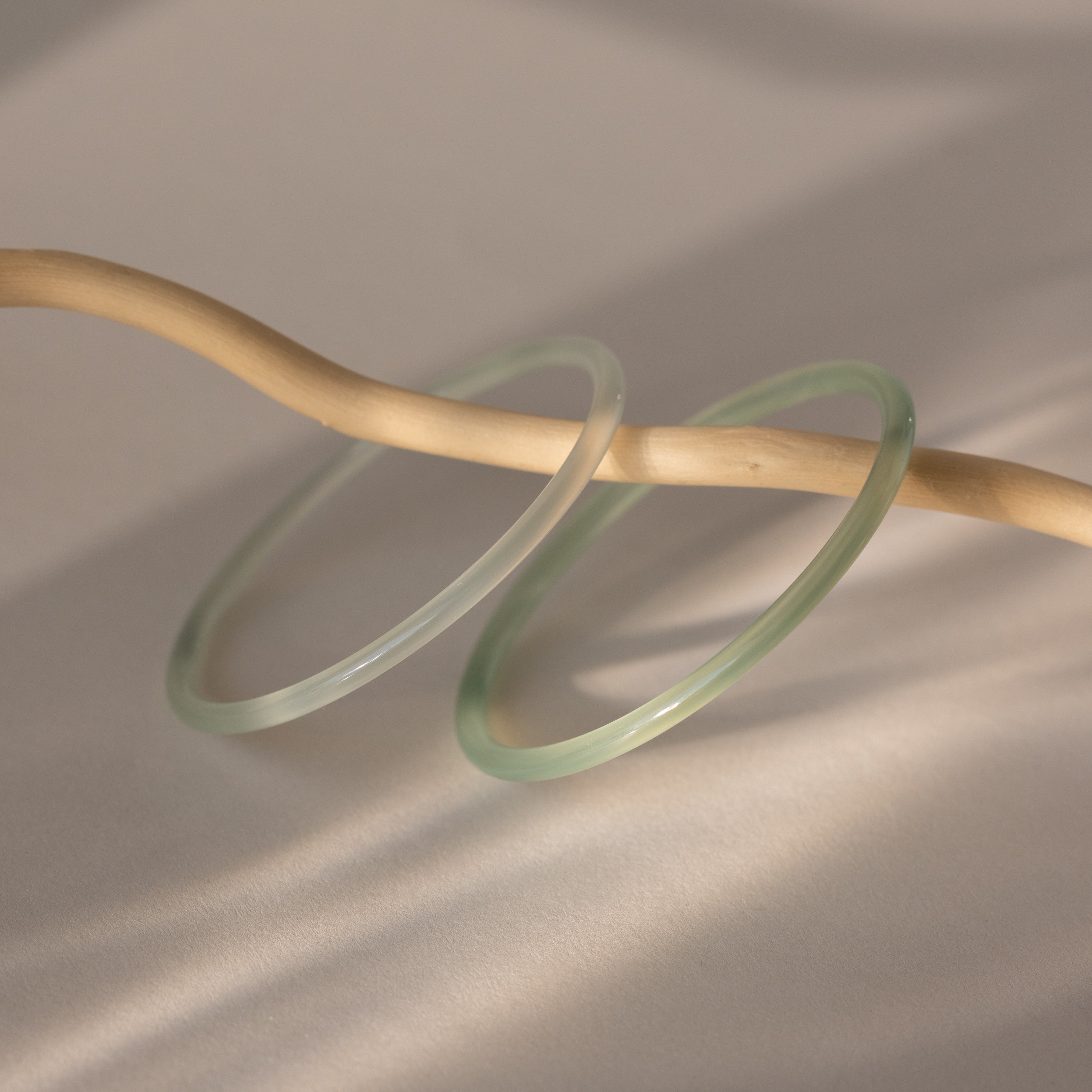 The Thin Jade Bangle Set, featuring two translucent green bangles with a glass-like finish, is displayed draped over a curved beige stick on a light neutral background.