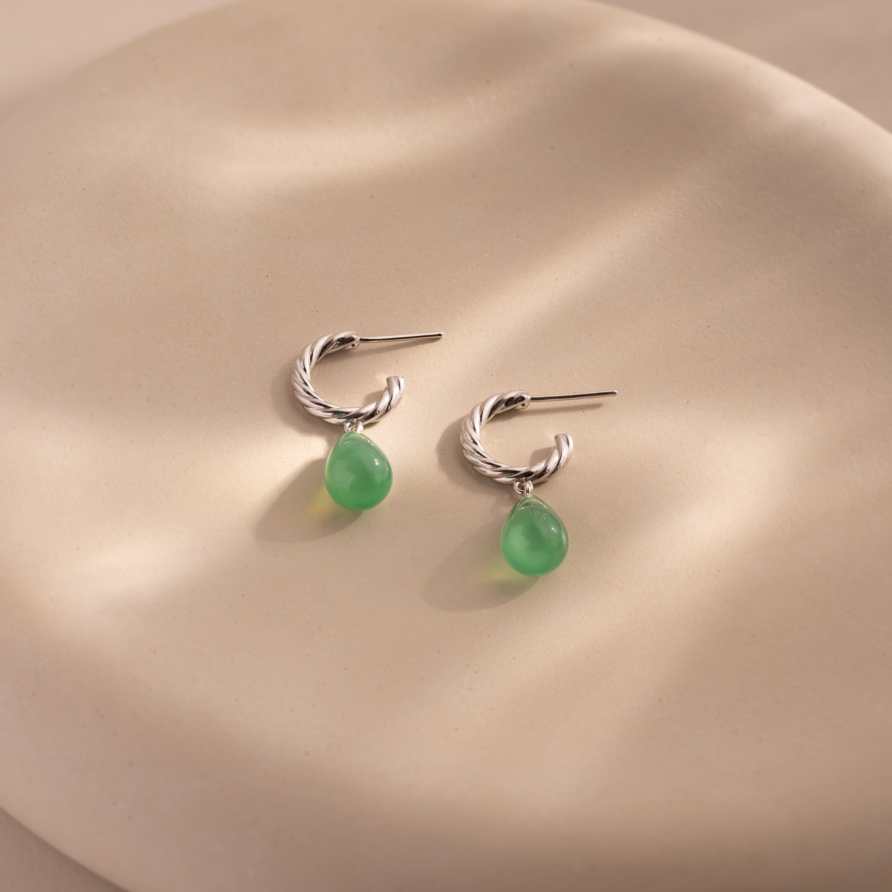 Twisted silver hoops with green jade drops elegantly hanging from a white ceramic vase.