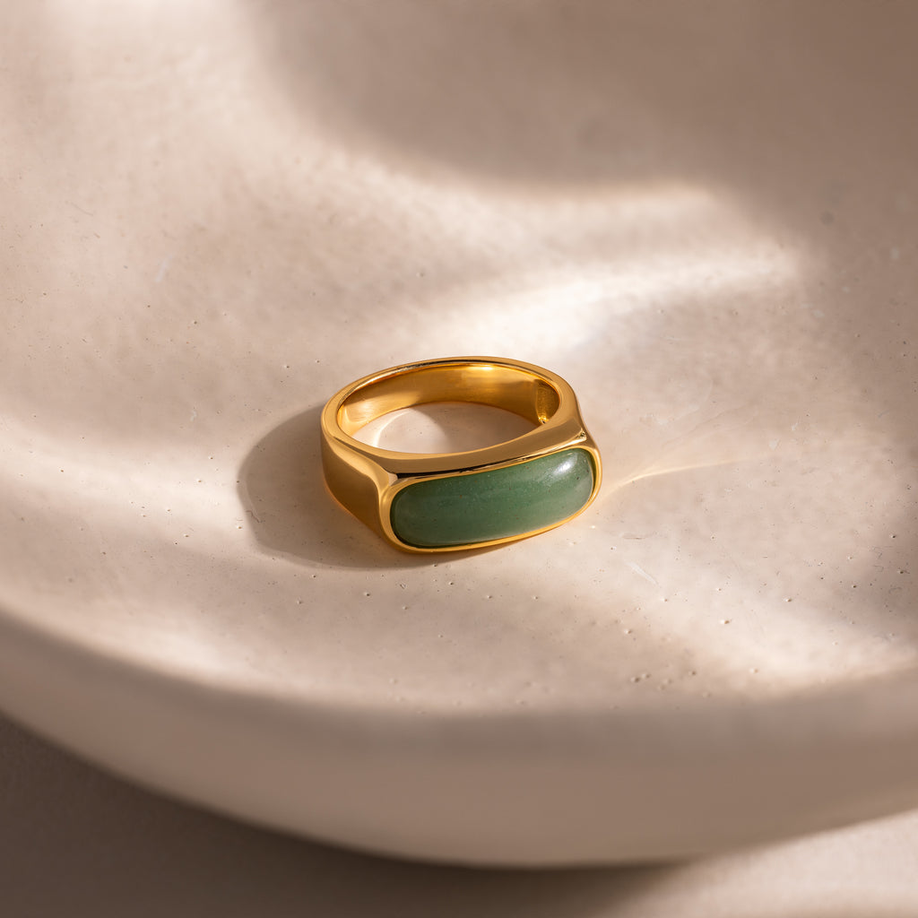 Close-up of a gold signet ring with a polished green jade gemstone inlay, displayed on a smooth ceramic dish.