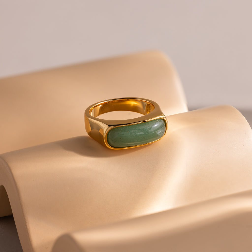 Close-up of a gold signet ring with a polished green jade gemstone inlay, displayed on a tan, wavy surface.