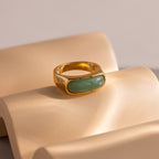 Close-up of a gold signet ring with a polished green jade gemstone inlay, displayed on a tan, wavy surface.
