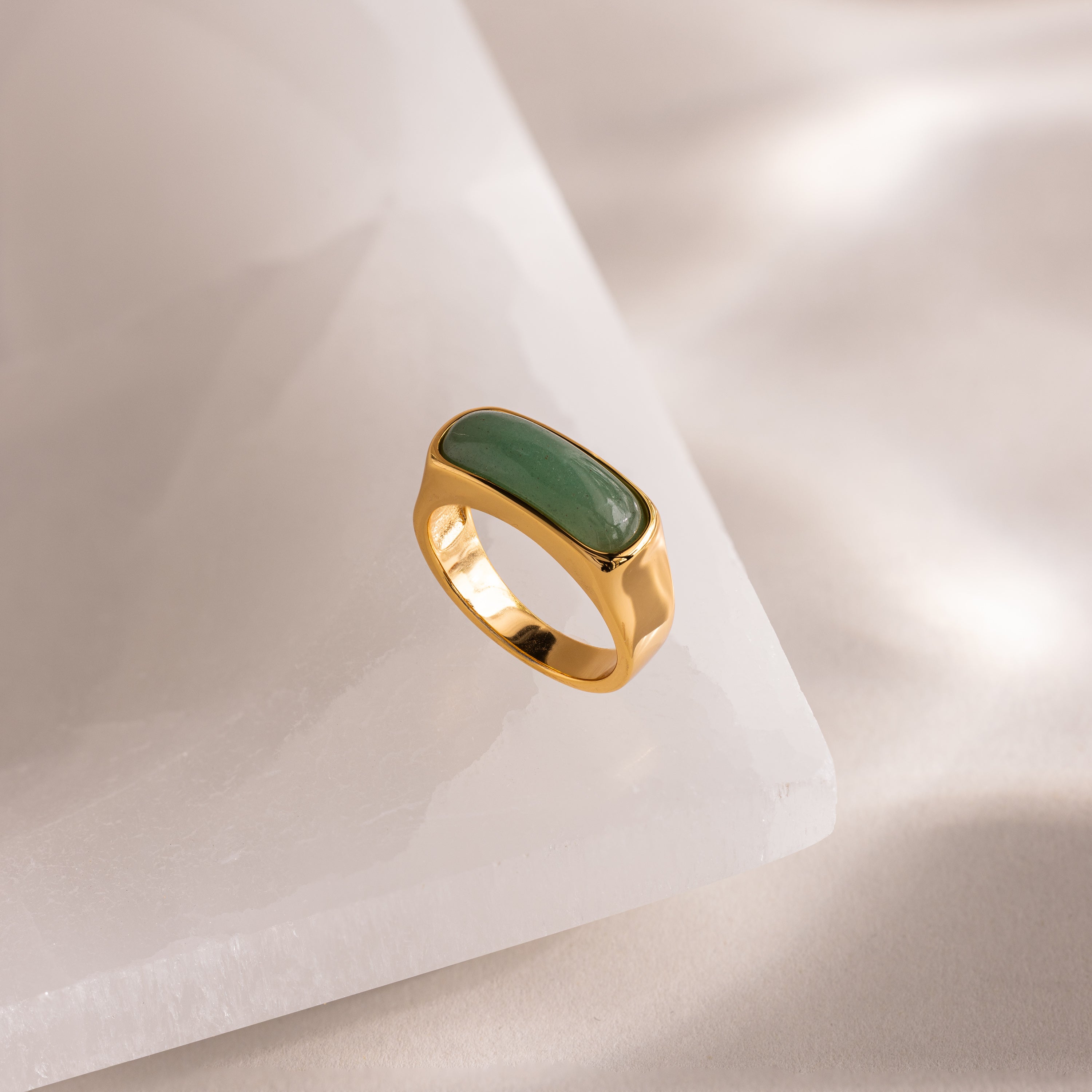 Close-up of a gold signet ring with a polished green jade gemstone inlay, positioned upright on a white stone surface.