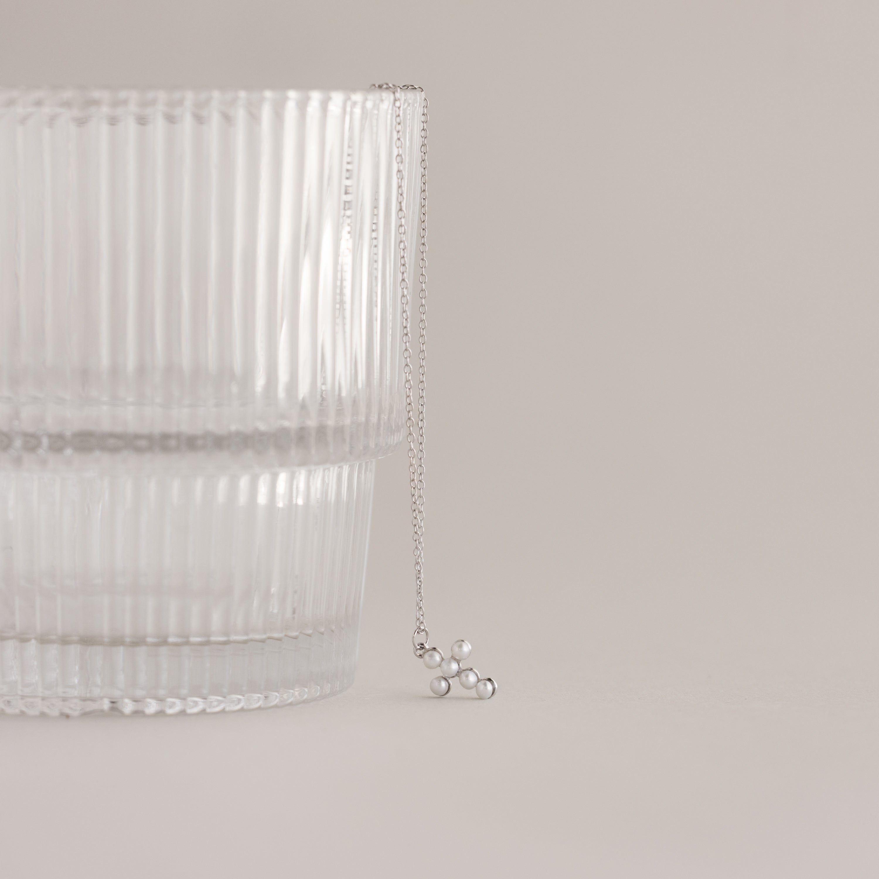 The Pearl Cross Necklace in Silver, featuring a delicate pendant, is displayed on a clear ribbed glass against a neutral background.