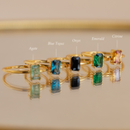 Five Emerald Cut Solitaire Rings with agate, blue topaz, onyx, emerald, and citrine stones rest on a reflective surface—each a vibrant birthstone ring that adds elegant style to any outfit.