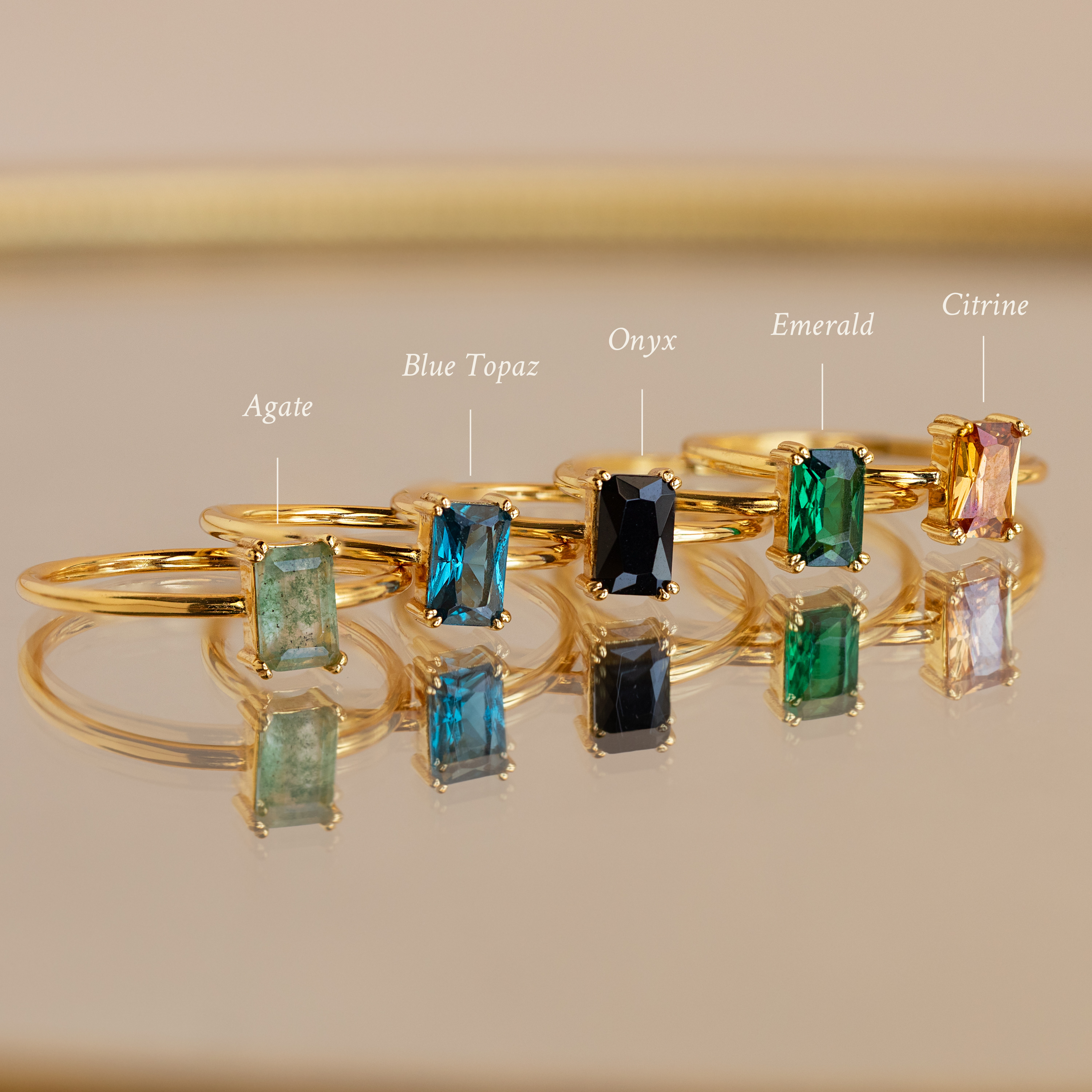 Five Emerald Cut Solitaire Rings with agate, blue topaz, onyx, emerald, and citrine stones rest on a reflective surface—each a vibrant birthstone ring that adds elegant style to any outfit.