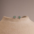 Close-up of silver flower-shaped stud earrings with a round green jade gemstone center, displayed on a neutral stone surface.