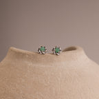 Close-up of silver flower-shaped stud earrings with a round green jade gemstone center, displayed on a neutral stone surface.