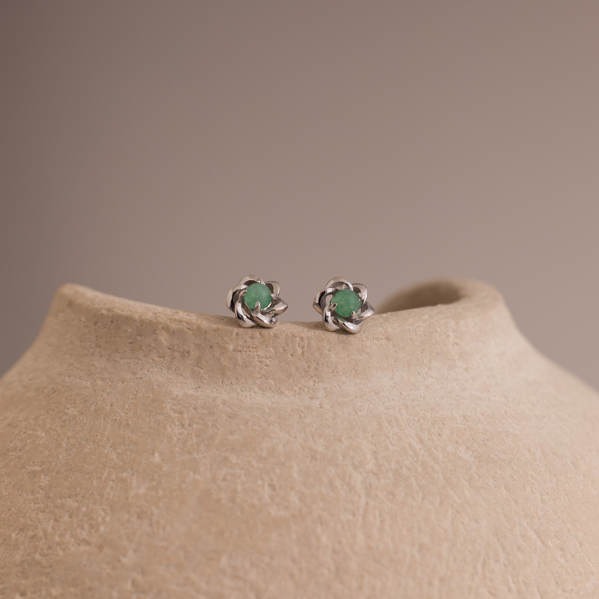 Close-up of silver flower-shaped stud earrings with a round green jade gemstone center, displayed on a neutral stone surface.