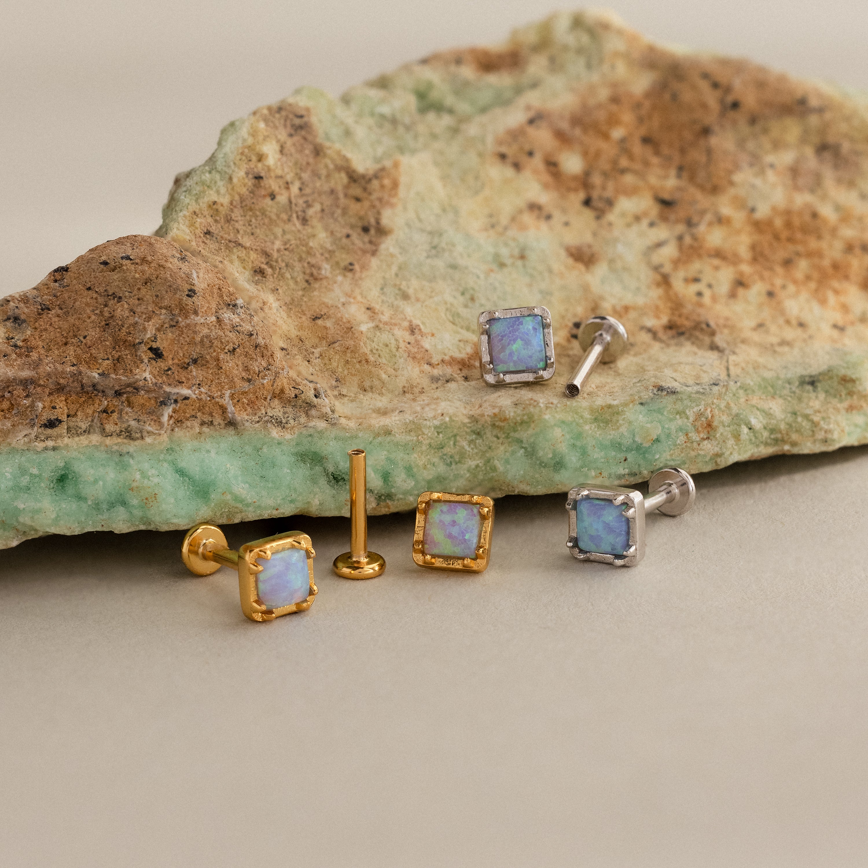 Blue Opal Prism Studs: Three pairs of square blue opal earrings in gold and silver, featuring iridescent stones, displayed on a textured rock.