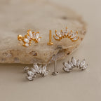 The Marquise Diamond Arc Climbers are marquise-cut crystal earrings in gold and silver, showcased on a textured beige stone surface.