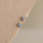 Blue Opal Prism Studs in Silver are displayed on a textured beige surface, reflecting the captivating charm of iridescent jewelry with every glance.