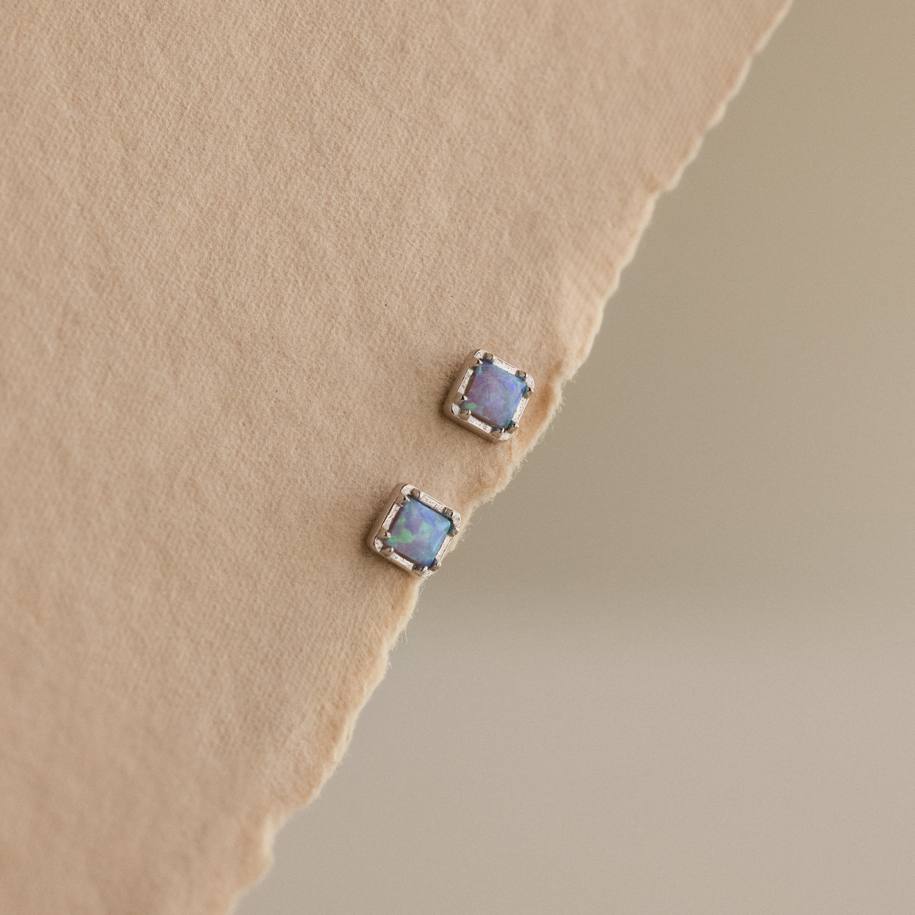 Blue Opal Prism Studs in Silver are displayed on a textured beige surface, reflecting the captivating charm of iridescent jewelry with every glance.