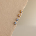 Blue Opal Prism Studs—two gold and two silver square earrings—are arranged on a textured beige surface, perfect for adding iridescent style to your look.