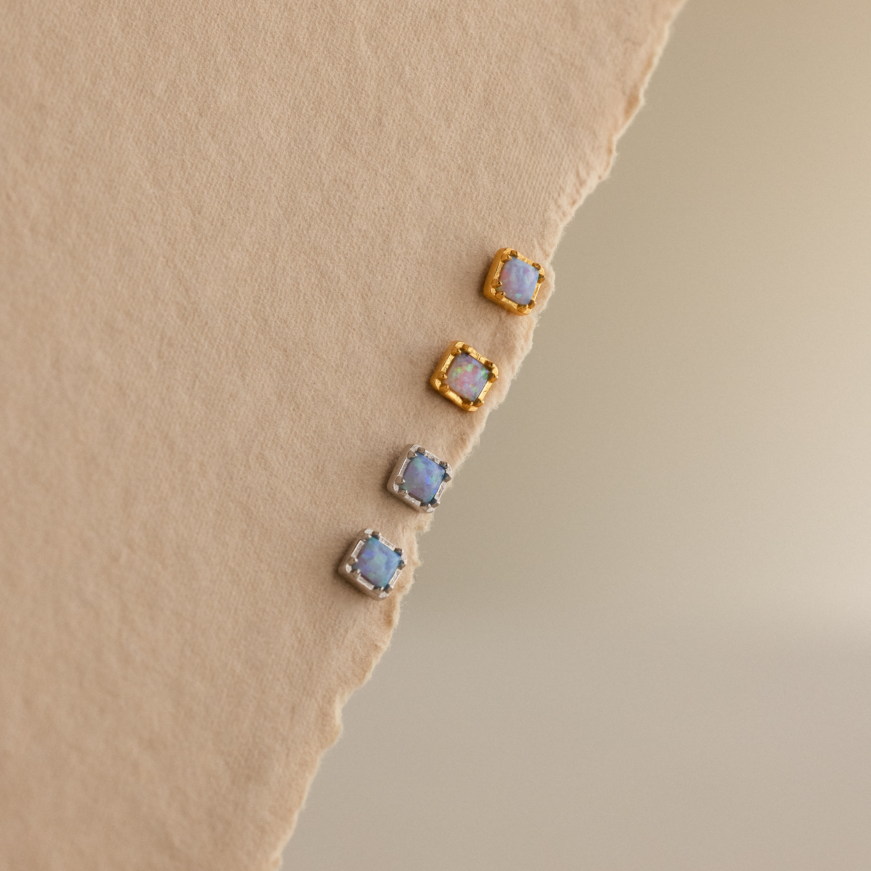 Blue Opal Prism Studs—two gold and two silver square earrings—are arranged on a textured beige surface, perfect for adding iridescent style to your look.