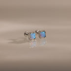 Blue Opal Prism Studs in Silver feature square blue opal gemstones that shimmer on a smooth, light beige surface—an elegant, iridescent touch for everyday wear.