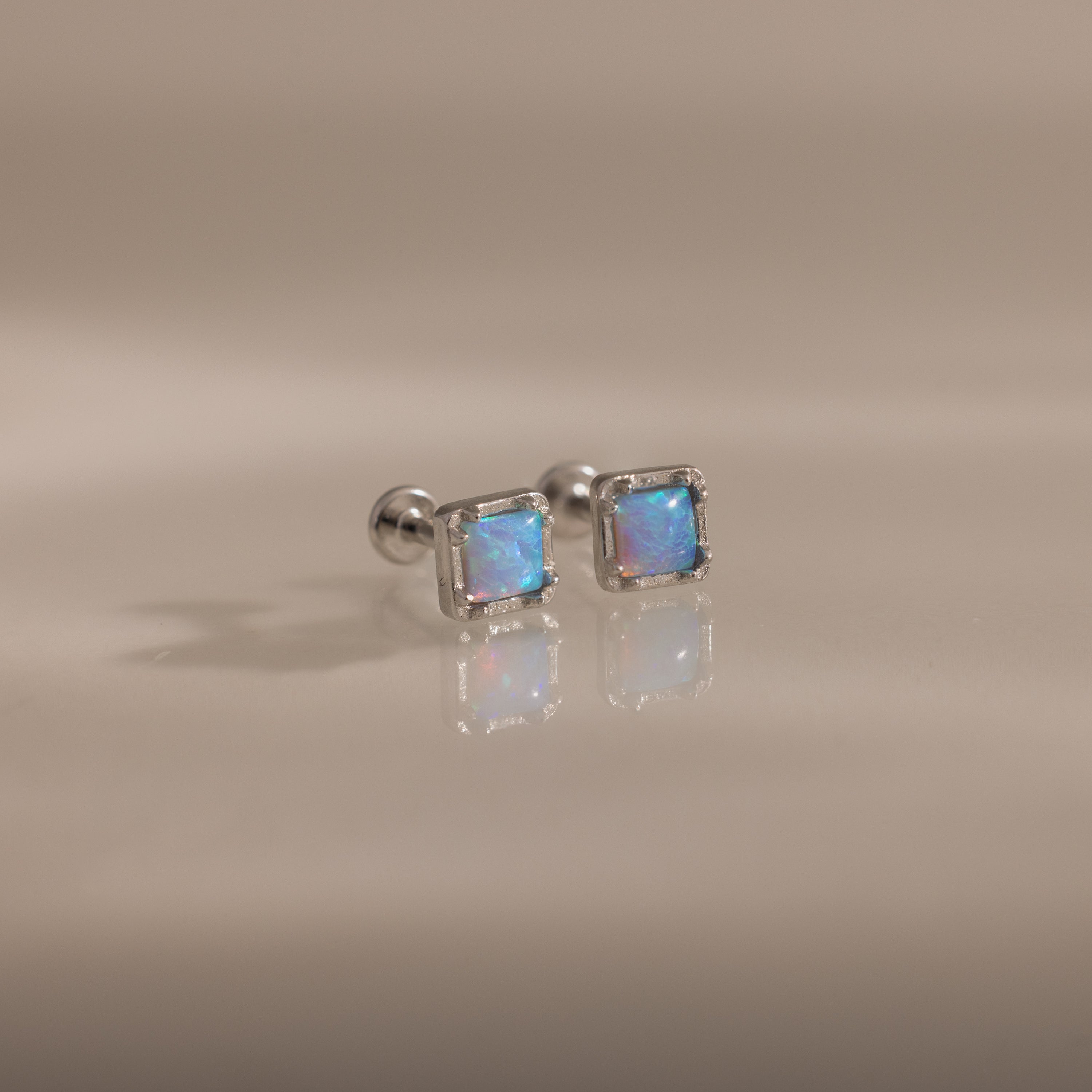 Blue Opal Prism Studs in Silver feature square blue opal gemstones that shimmer on a smooth, light beige surface—an elegant, iridescent touch for everyday wear.