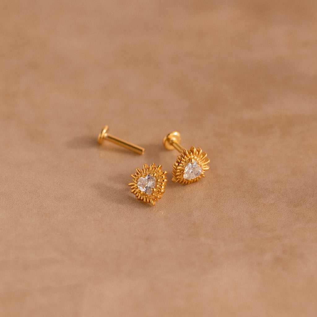 Sacred Heart Studs: gold earrings with clear gemstones and a delicate sunburst texture, displayed on a soft beige surface.