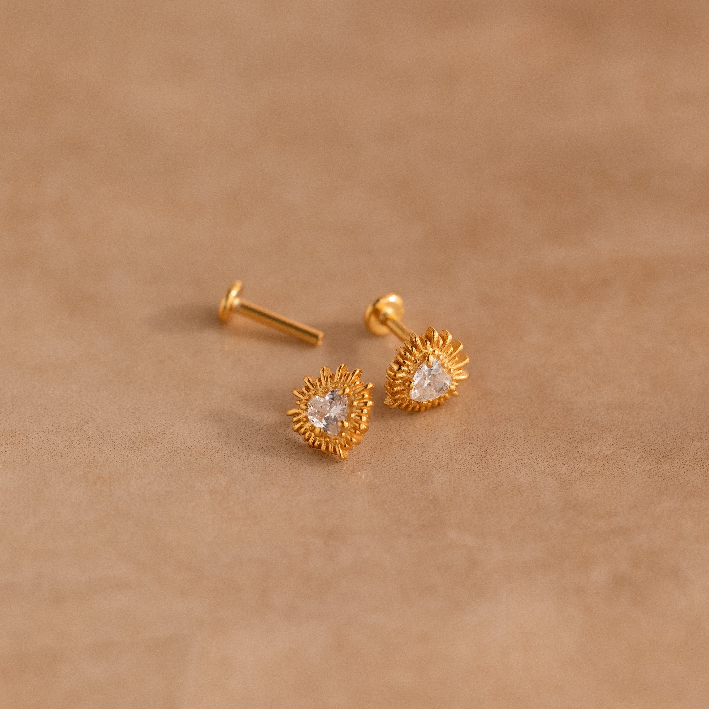 Sacred Heart Studs: gold earrings with clear gemstones and a delicate sunburst texture, displayed on a soft beige surface.