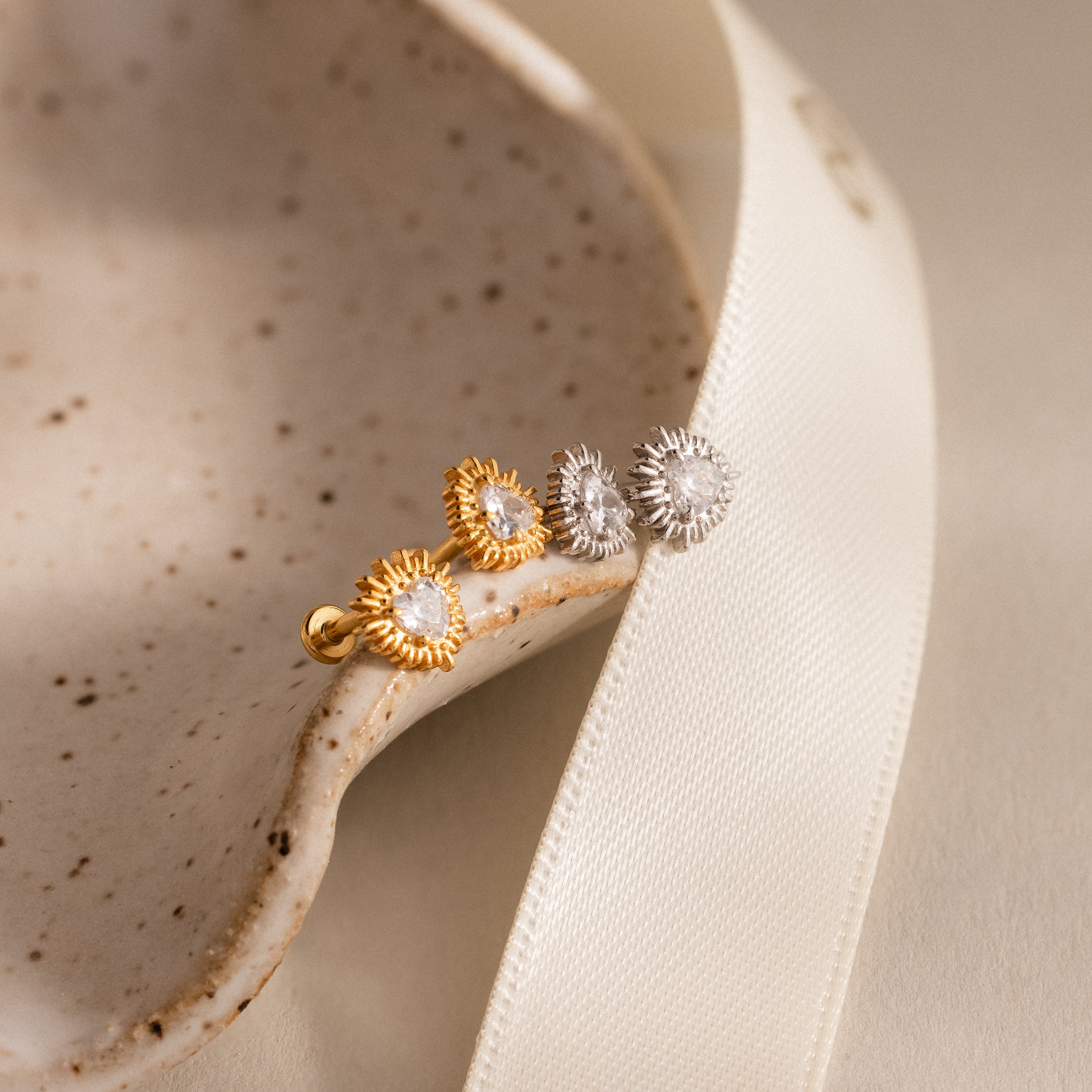 Sacred Heart Studs: gold and silver sunburst earrings with clear stones, featuring a striking sunburst texture, displayed on a speckled dish and cream ribbon.