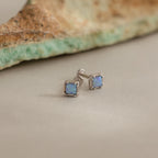 Blue Opal Prism Studs in Silver are displayed on a neutral surface, highlighting their iridescent charm, with a matching stone featured in the background.
