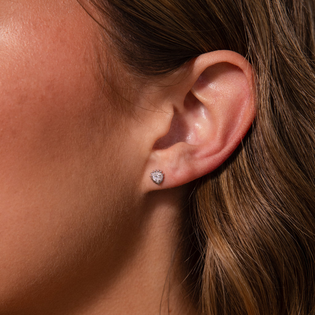 Close-up of a womans ear wearing Sacred Heart Studs in Silver—small, pear-shaped earrings with a subtle sunburst texture.