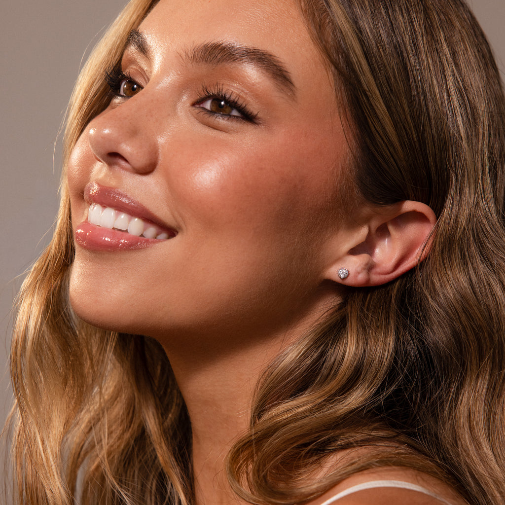 Woman smiling with long wavy hair and glowing skin, wearing Sacred Heart Studs in Silver featuring a subtle sunburst texture, facing left.