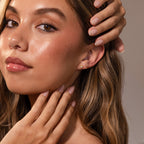 A woman with long brown hair and natural makeup touches her face while wearing Sacred Heart Studs, featuring a sunburst texture.