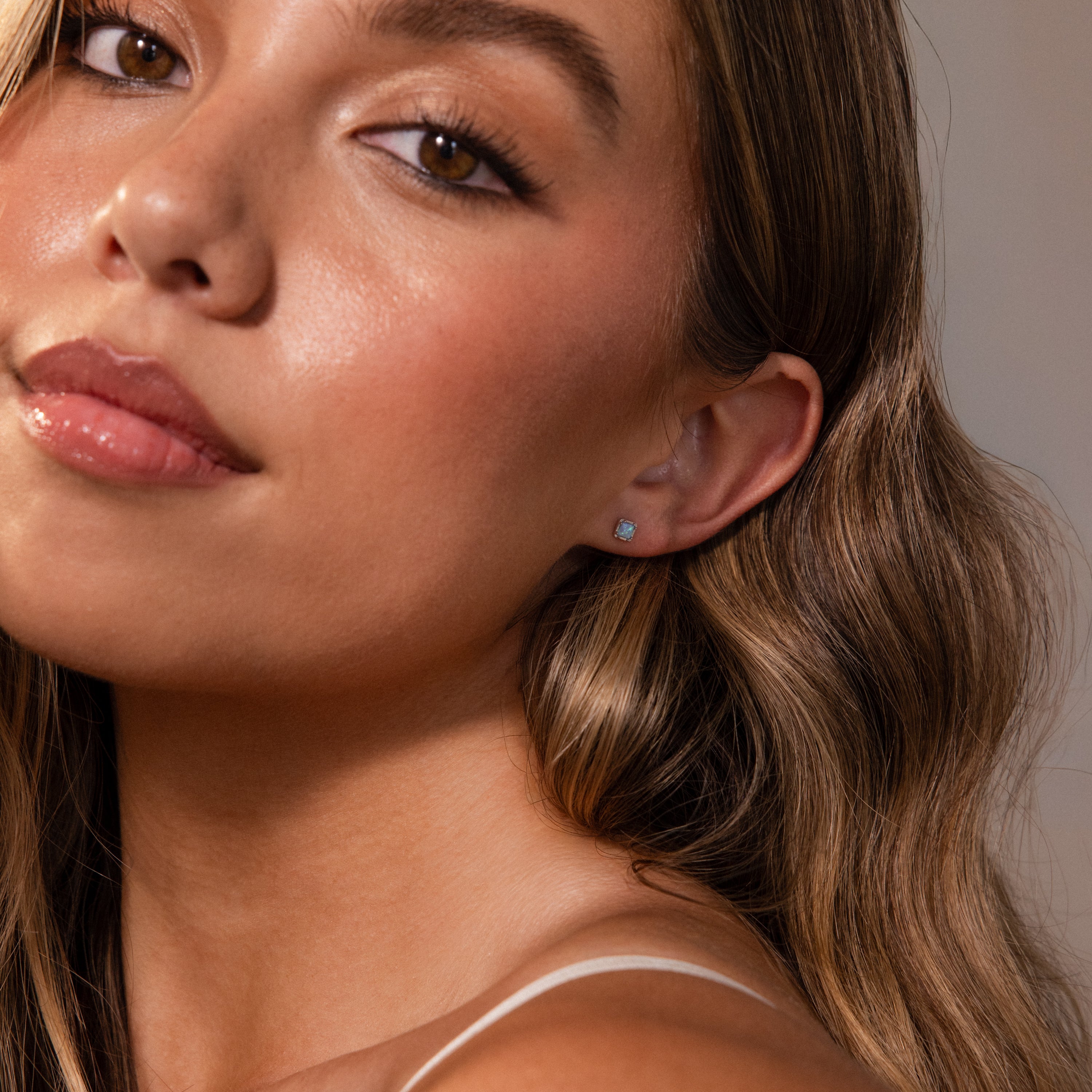 A close-up of a young woman with wavy brown hair, glowing skin, and delicate Blue Opal Prism Studs in Silver that add a touch of iridescent beauty.