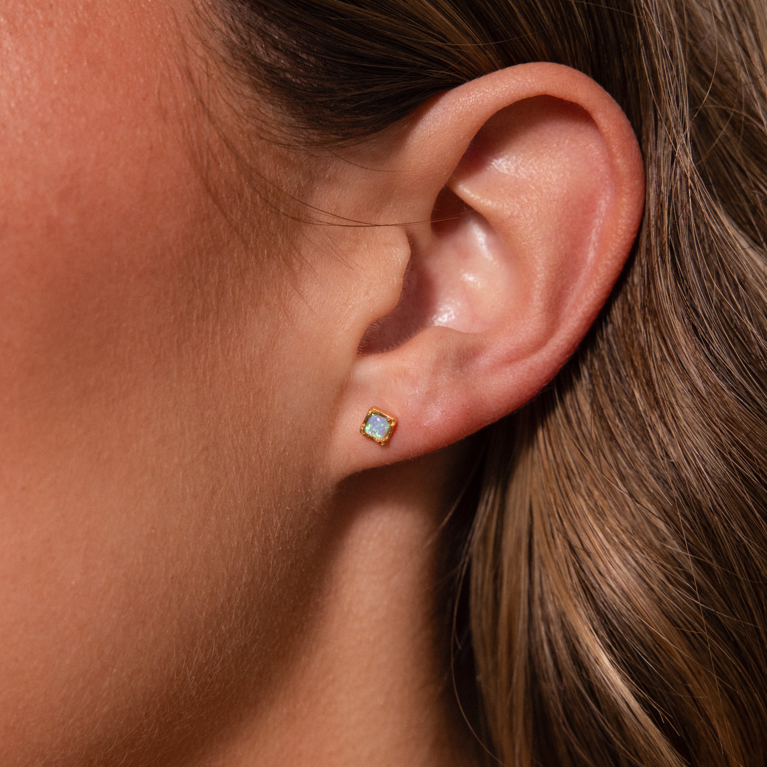 Close-up of a womans ear wearing Blue Opal Prism Studs with a gold setting, highlighting the earrings iridescent shimmer and elegant square design.