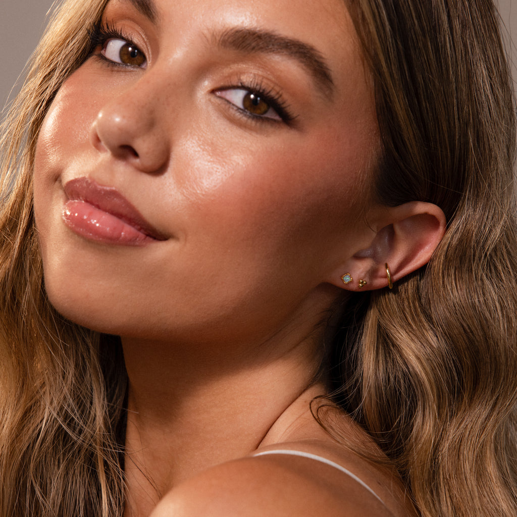 Woman with long wavy hair, glowing skin, and Blue Opal Prism Studs, looking over her shoulder and smiling softly.