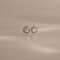 The Sacred Heart Studs in Silver feature round sparkling stones and a sunburst texture, shown on a smooth beige surface.