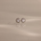 The Sacred Heart Studs in Silver feature round sparkling stones and a sunburst texture, shown on a smooth beige surface.