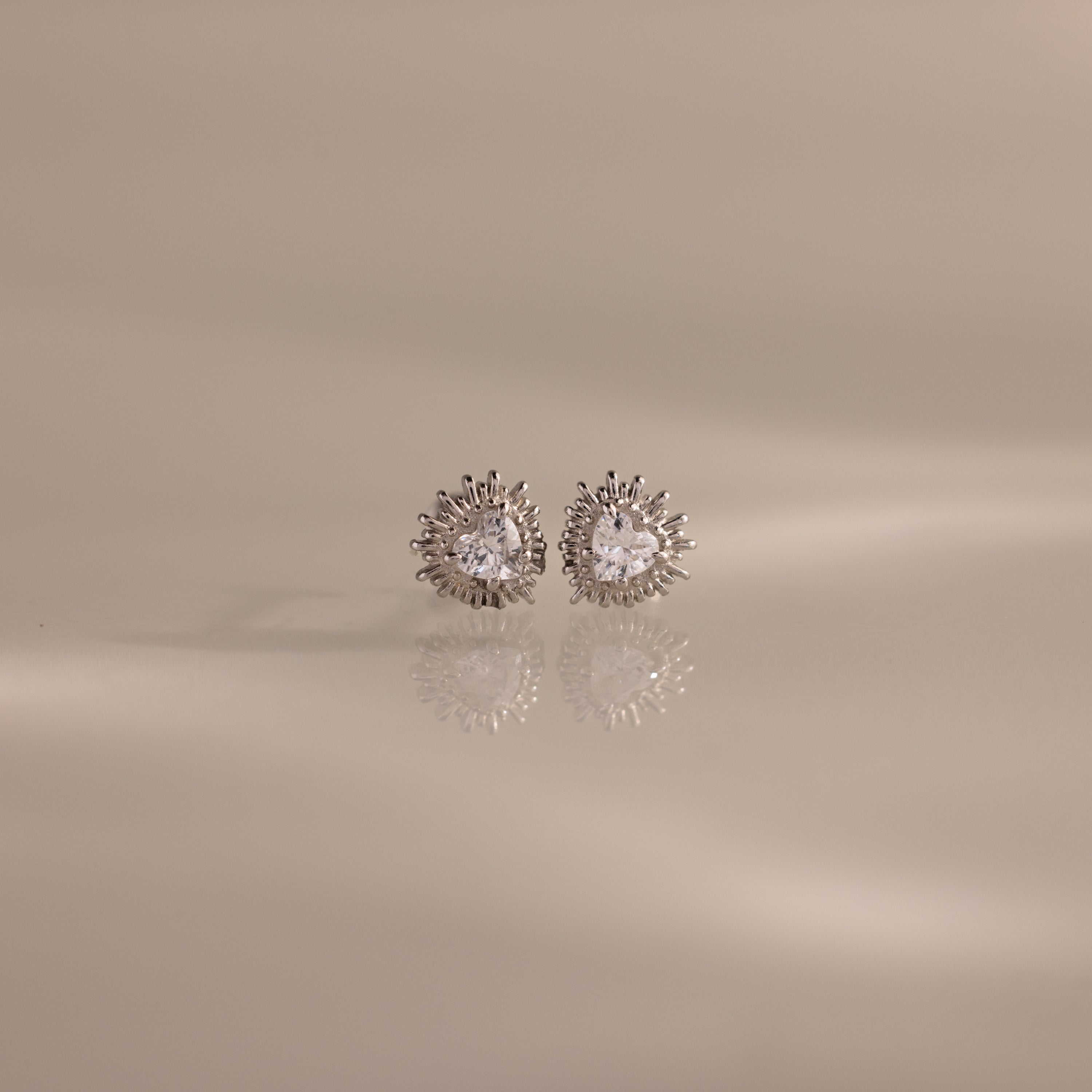 The Sacred Heart Studs in Silver feature round sparkling stones and a sunburst texture, shown on a smooth beige surface.