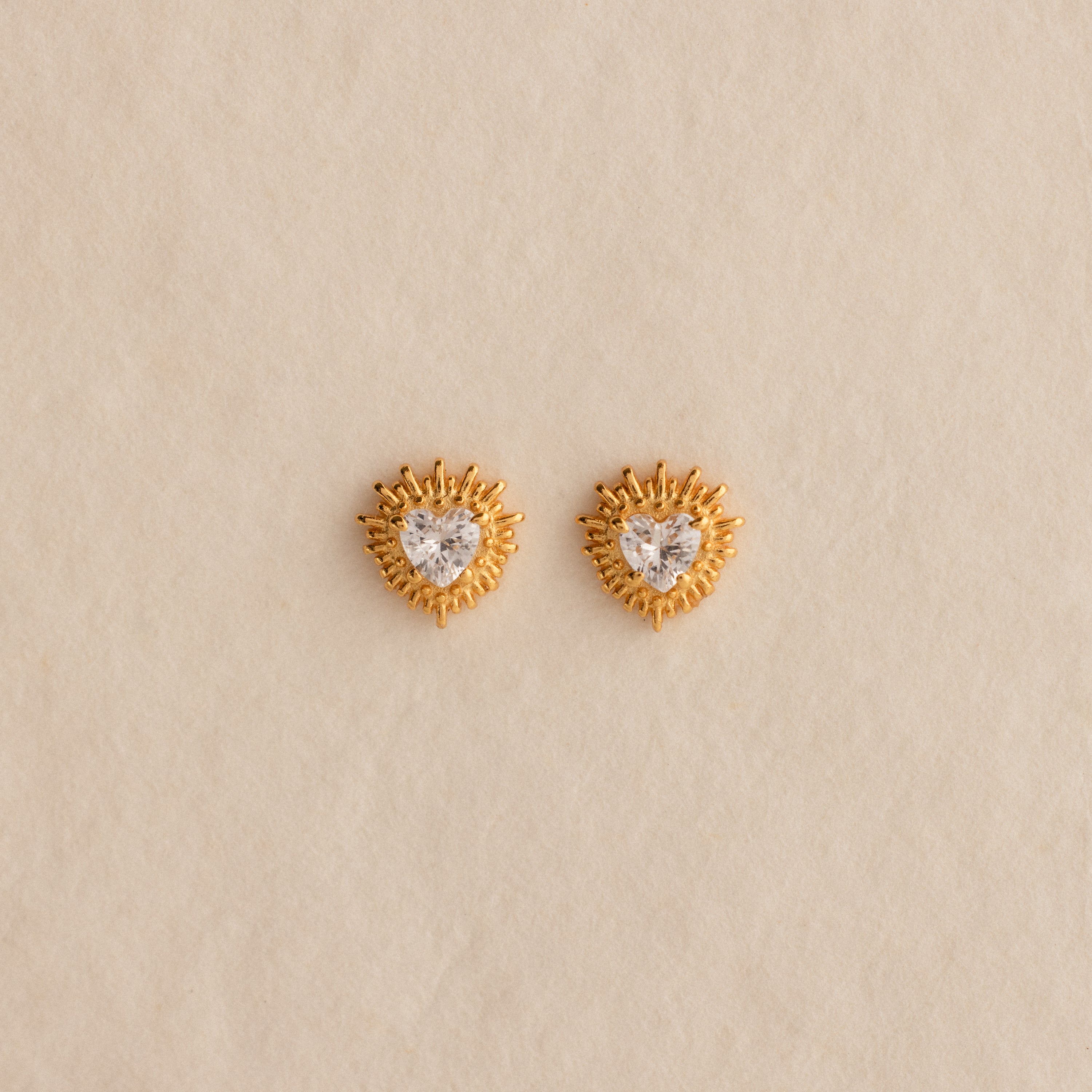Sacred Heart Studs in 18K Gold, featuring a heart-shaped center stone and radiant sunburst texture, displayed on a beige background.