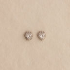 The Sacred Heart Studs in Silver are faceted heart earrings with clear stones, shown on a beige textured background.