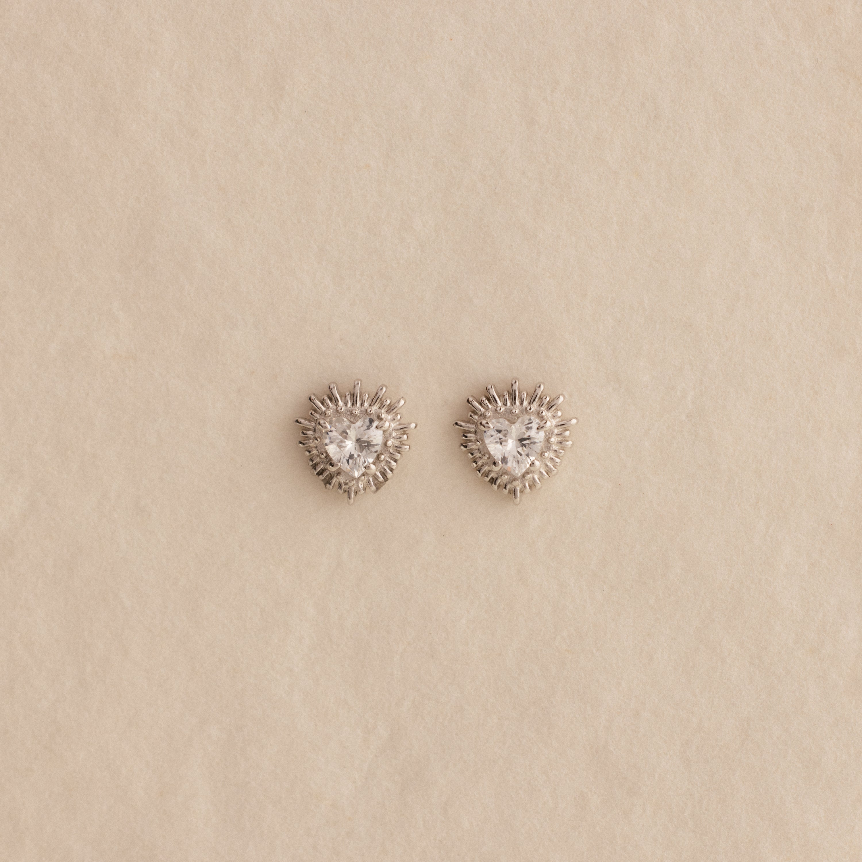 The Sacred Heart Studs in Silver are faceted heart earrings with clear stones, shown on a beige textured background.