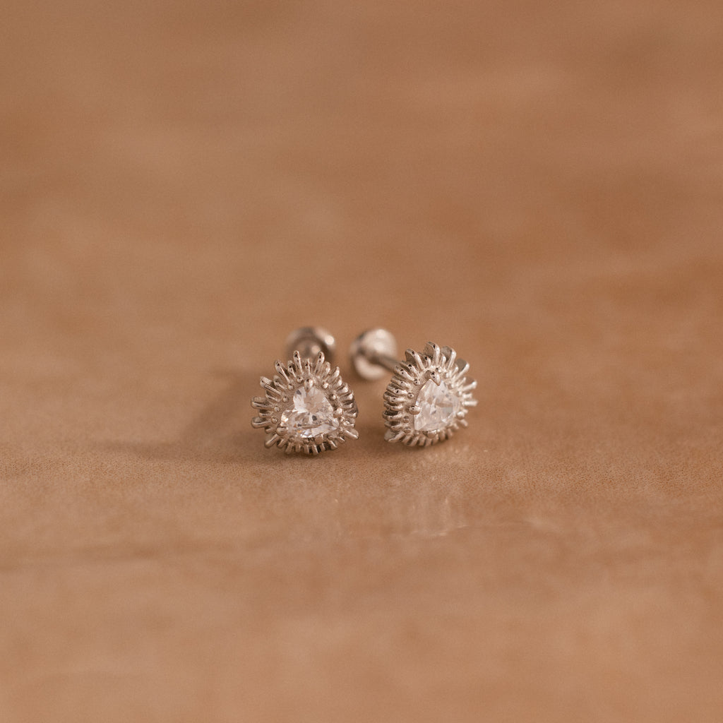 The Sacred Heart Studs in Silver, featuring teardrop-shaped clear stones and a sunburst texture, add a radiant touch as they rest on a soft brown surface.