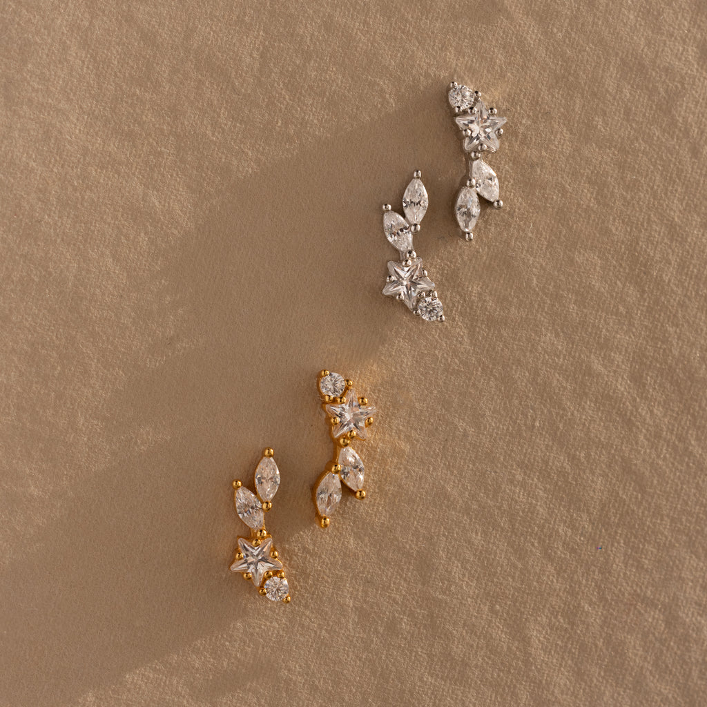 A pair of Diamond Starfall Climbers, gold earrings with star accents, displayed on a textured beige surface.
