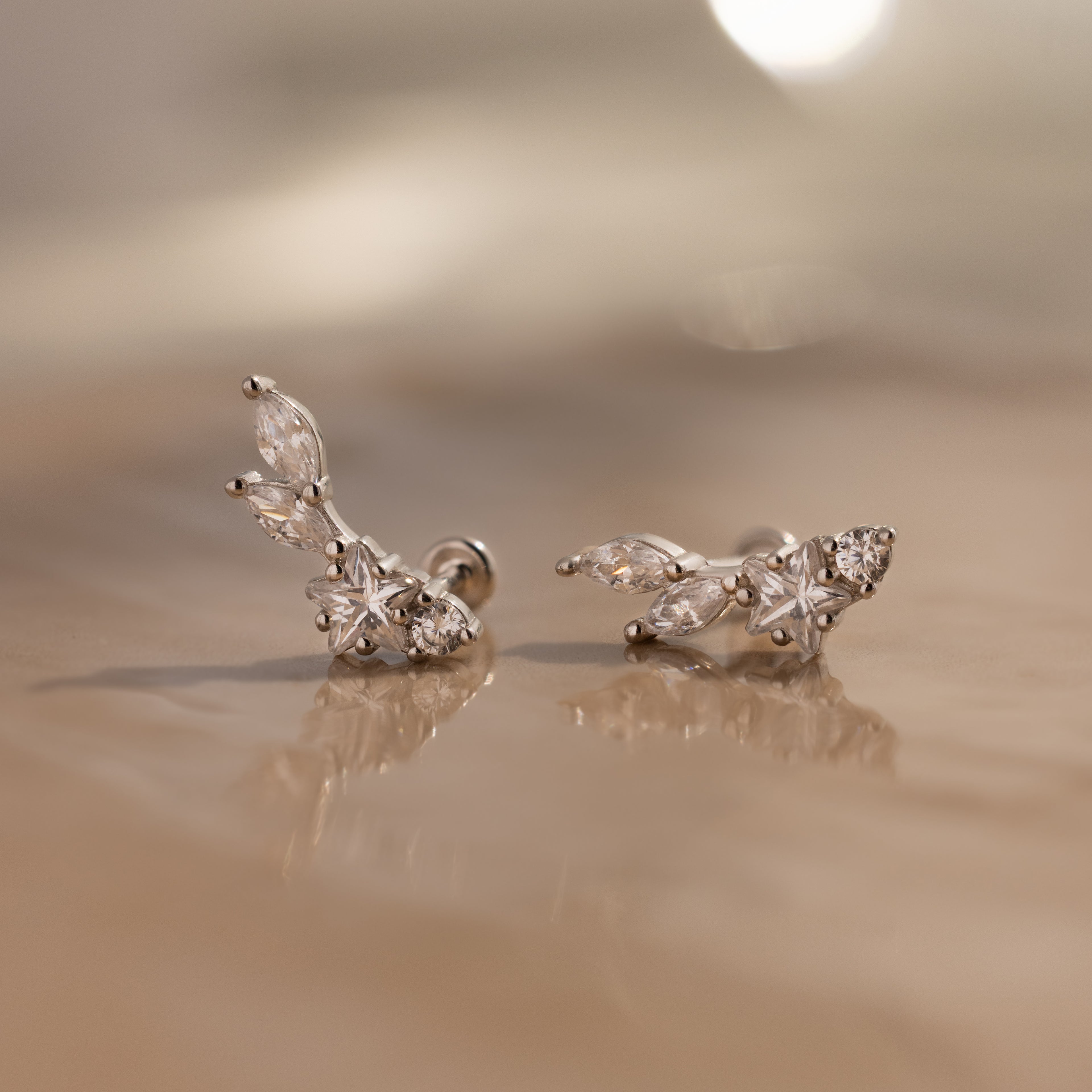 Diamond Starfall Climbers—delicate silver earrings with marquise-cut and round clear stones plus a star accent—rest on a reflective beige surface.