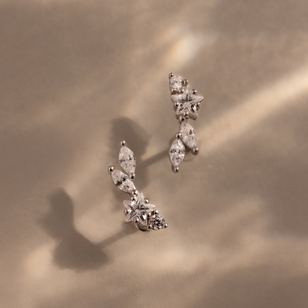 The Diamond Starfall Climbers in Silver, featuring marquise cuts with delicate leaf and star designs, rest on a beige surface bathed in soft light and shadows for an elegant effect.