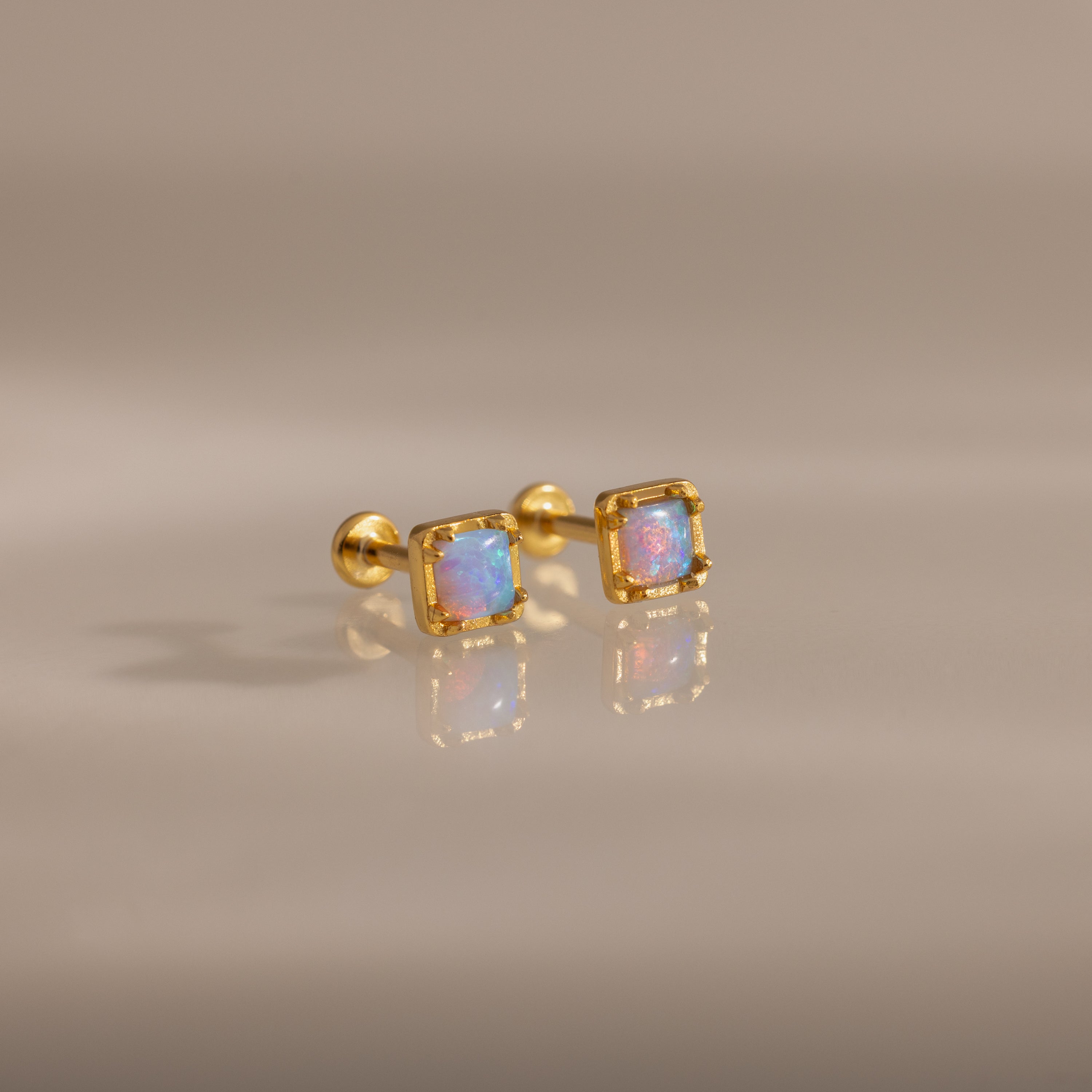 Blue Opal Prism Studs featuring square blue opal gems, displayed as iridescent jewelry on a reflective beige surface.