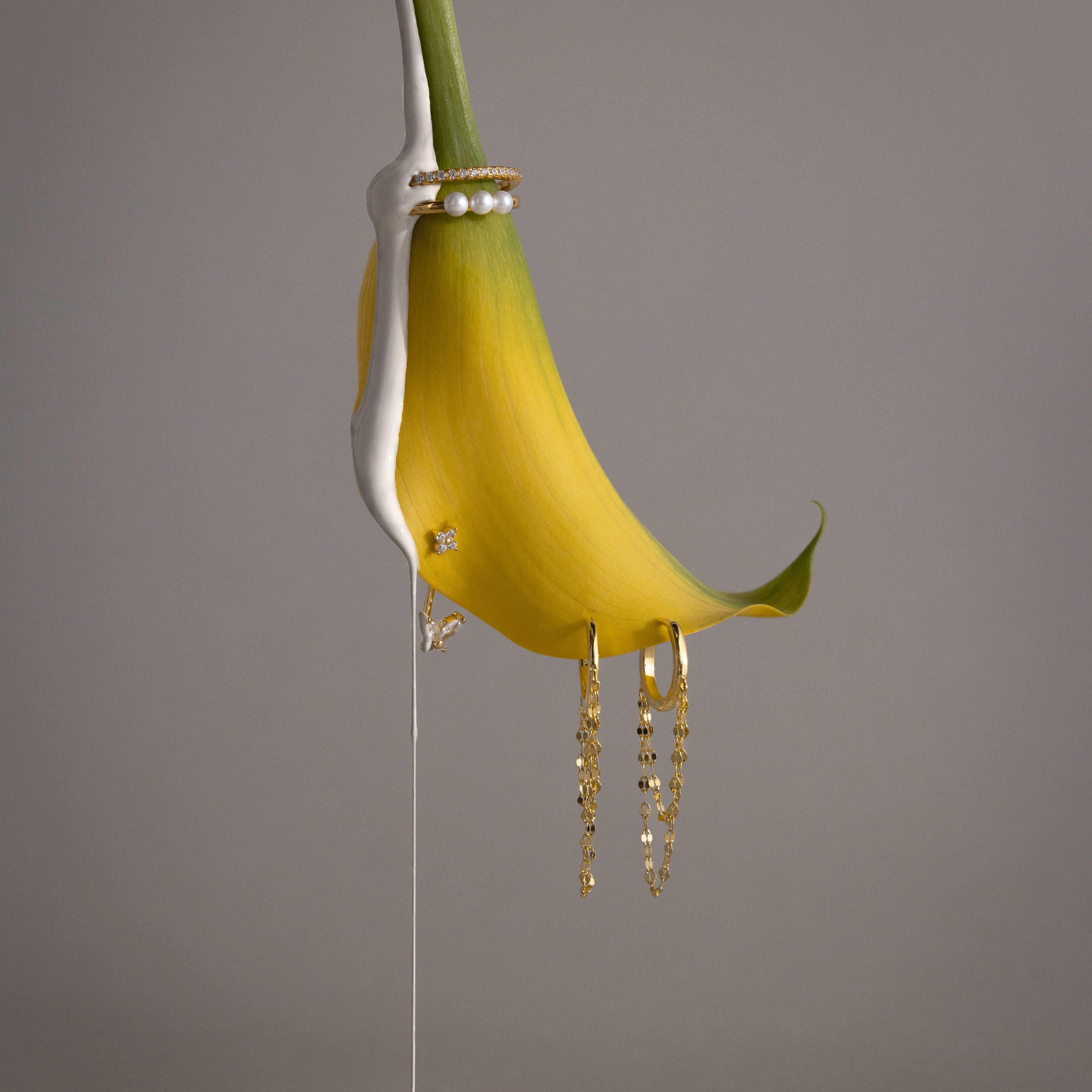 A yellow calla lily adorned with Butterfly Ear Jackets in 18K Gold and pearls, partially dipped in white paint, set against a gray background.