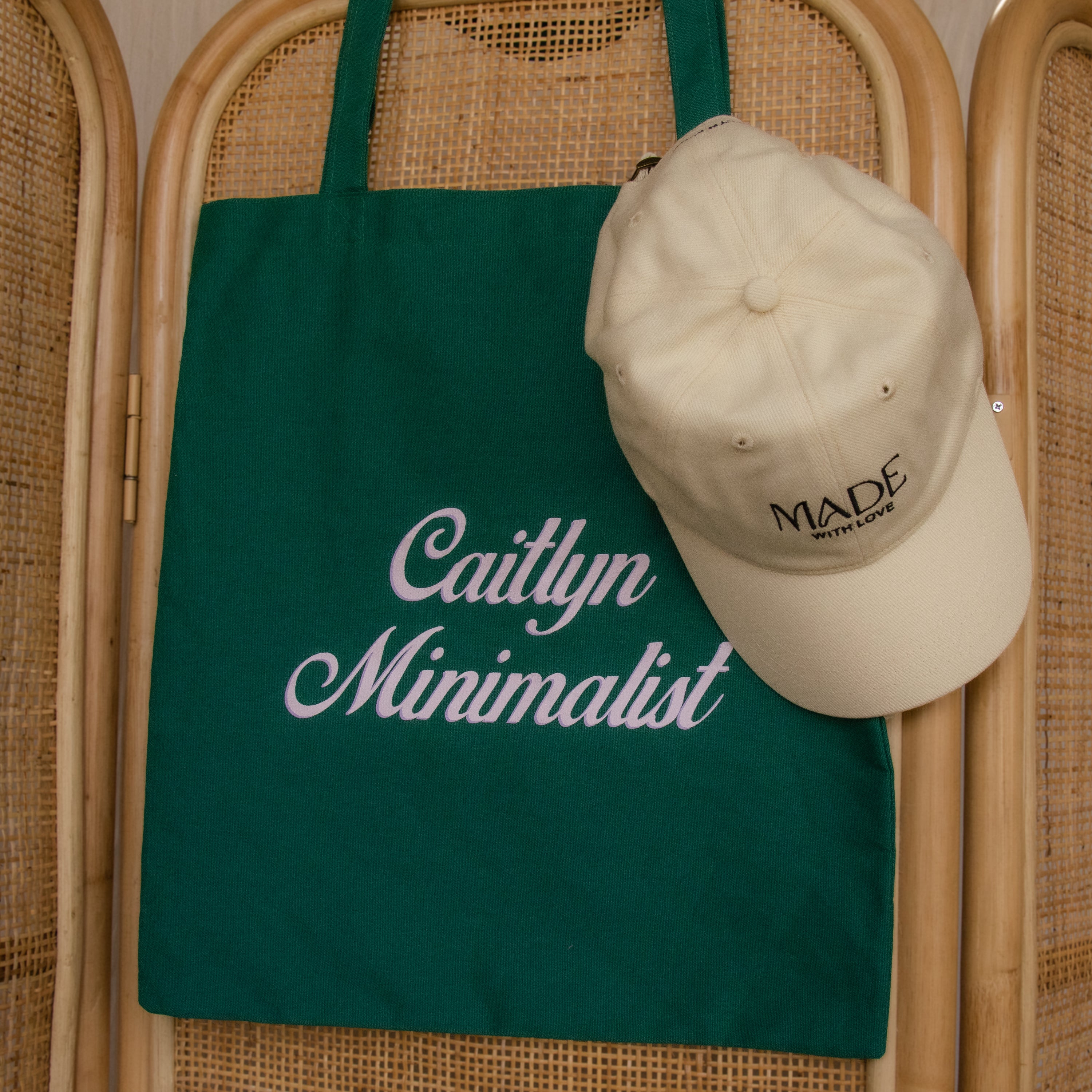Free Gift of Choice: A green tote bag with Caitlyn Minimalist text and a cream cap reading MADE WITH LOVE hang on a wooden chair—ideal for fans of stylish accessories.