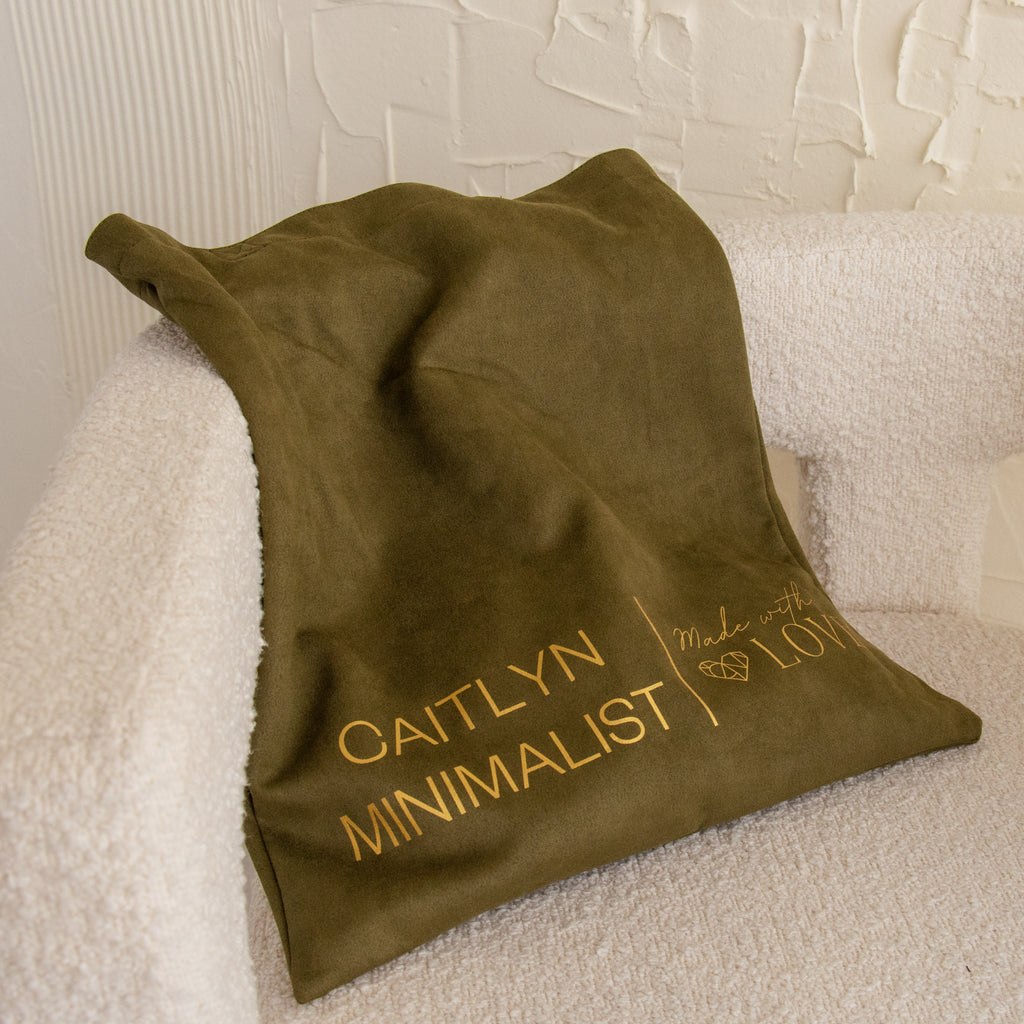 A cream chair is draped with an olive green cloth featuring “CAITLYN MINIMALIST Made with LOVE”—the perfect accent to pair with mugs or totes. Choose this as your Free Gift of Choice for a thoughtful touch.