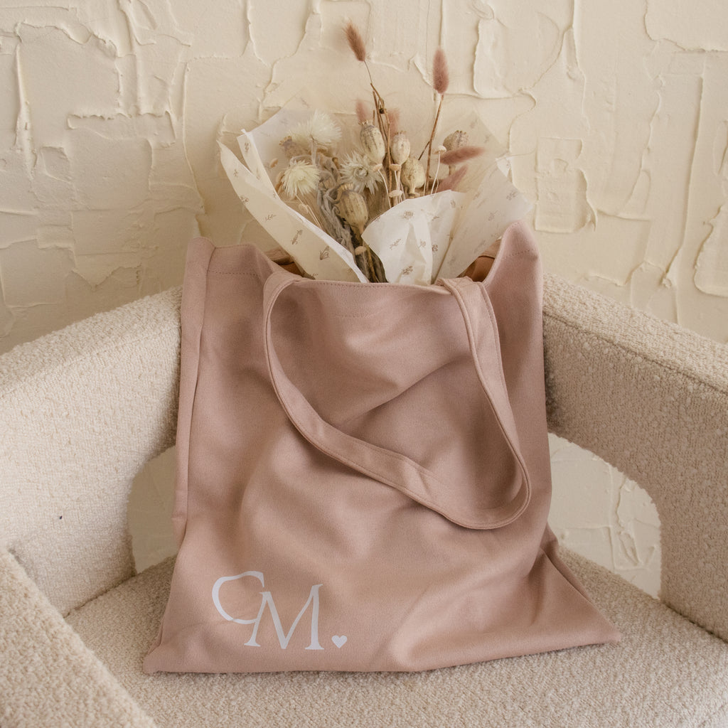 A beige tote bag with “CM.” initials, filled with dried flowers, rests on a cream chair—perfect for your essentials or as a Free Gift of Choice when you buy a cute mug.