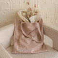 A beige tote bag with “CM.” initials, filled with dried flowers, rests on a cream chair—perfect for your essentials or as a Free Gift of Choice when you buy a cute mug.