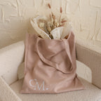 A beige tote bag with “CM.” initials, filled with dried flowers, rests on a cream chair—perfect for your essentials or as a Free Gift of Choice when you buy a cute mug.