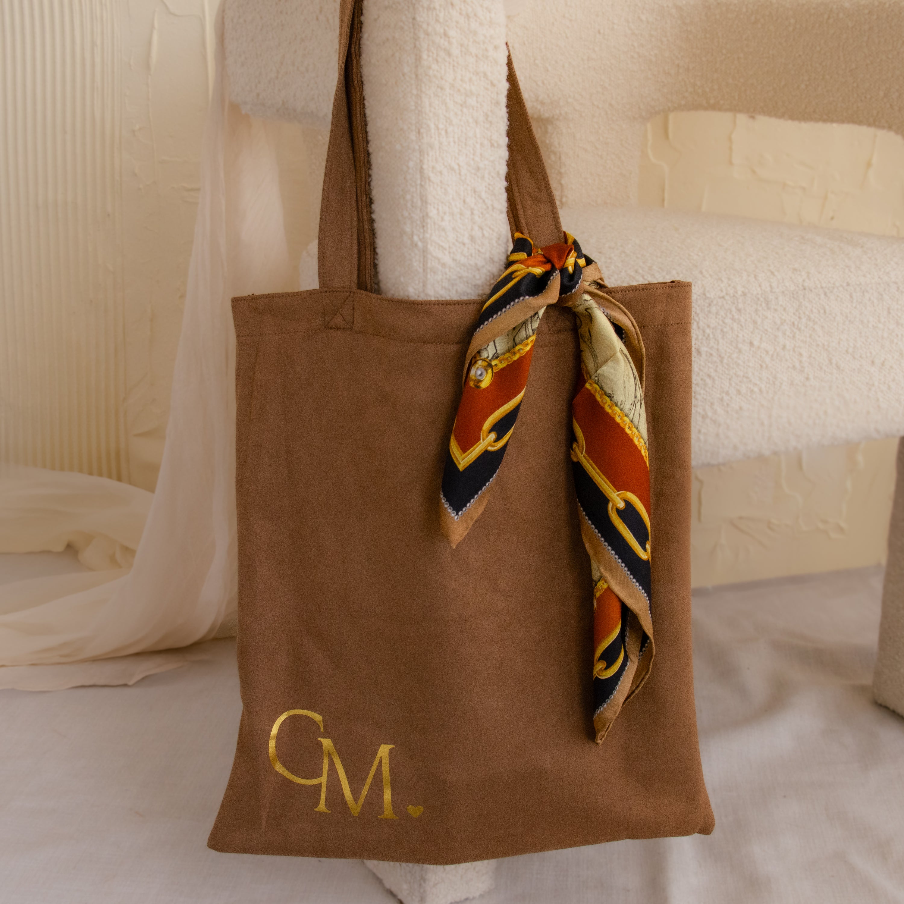 The Free Gift of Choice brown tote bag features gold initials "CM" and a patterned scarf on one handle—stylish for carrying your essentials or pairing with other cute totes.
