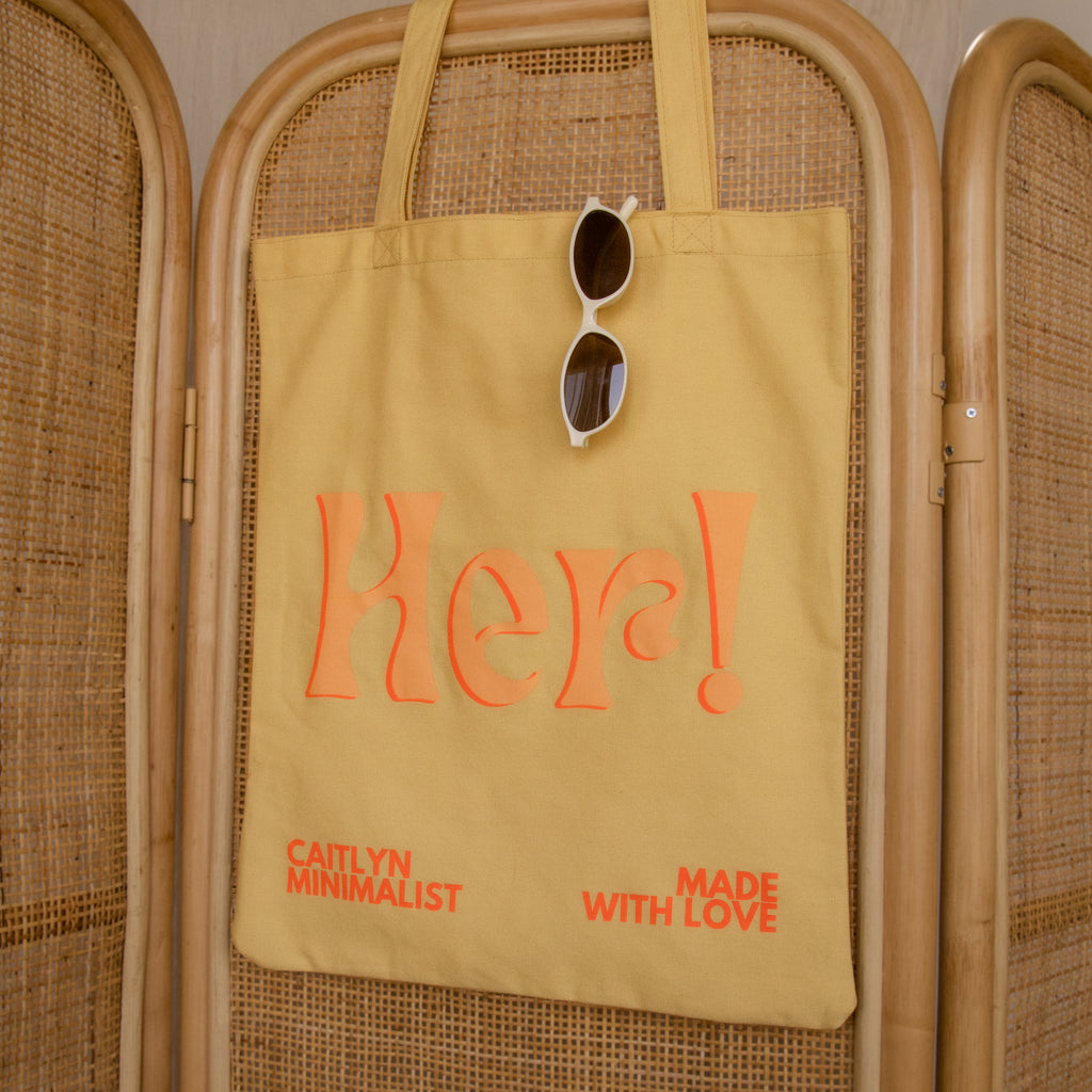 Brighten your day with the Free Gift of Choice—a yellow tote bag featuring "Her!" in orange text and sunglasses detail, making it a perfect pick for tote bag lovers or as a cheerful free gift.